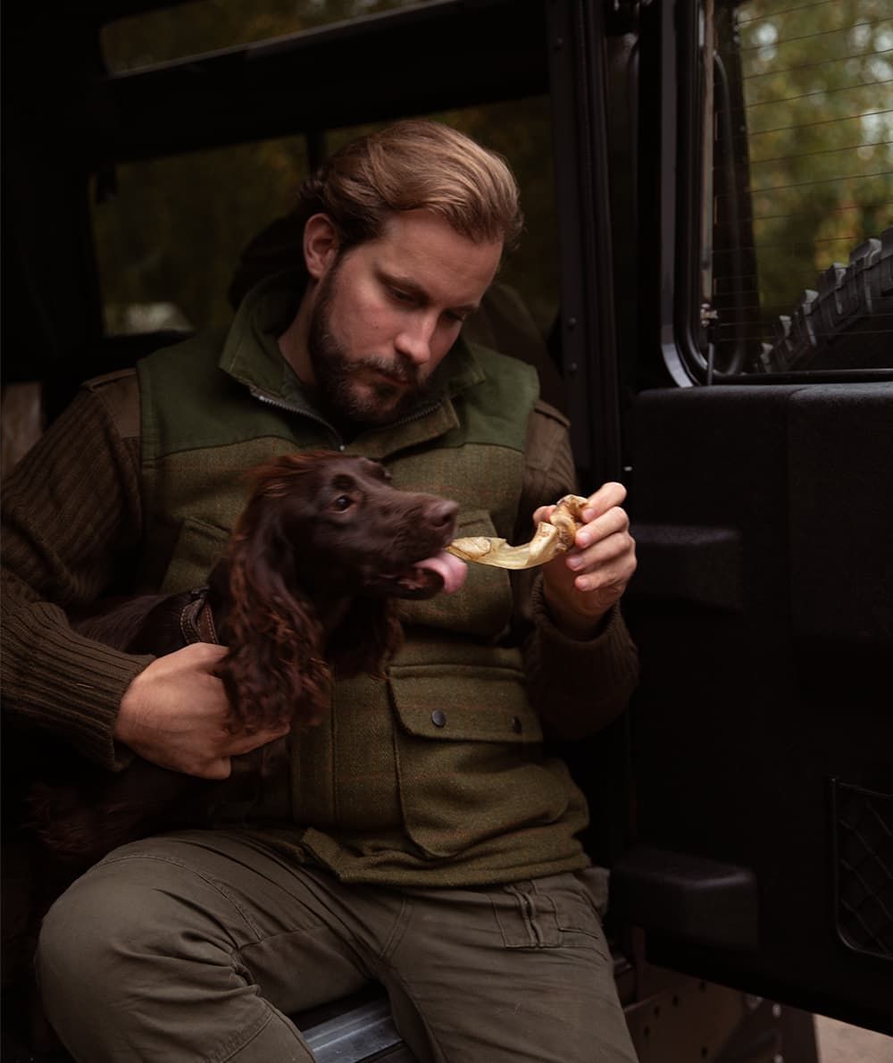 En mand med en hund sidder på bagsædet af et køretøj og nyder ESSENTIAL Cow Delights XL (okseører) 2 stk i posen - god tyggeaktivitet fra Essential.